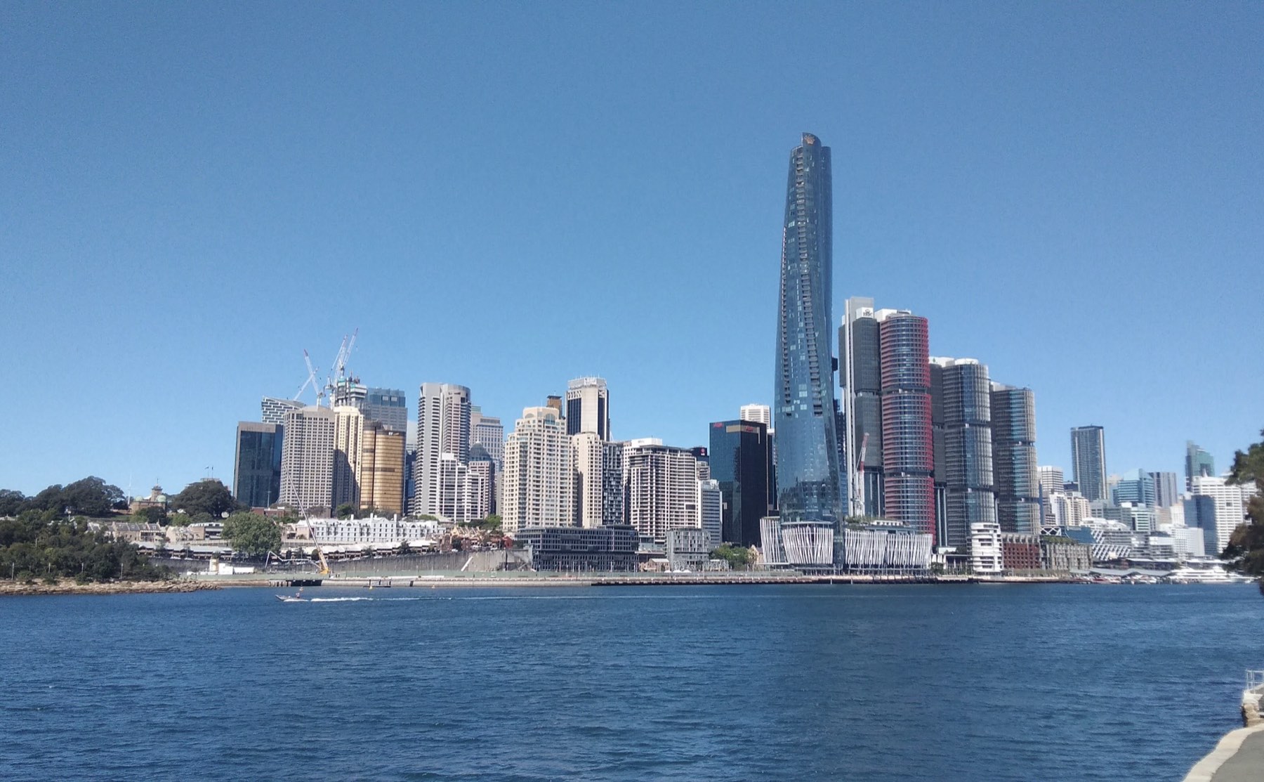 The tall, sculpted glass tower of Crown Sydney rising above Barangaroo and the Sydney skyline across the blue harbour water