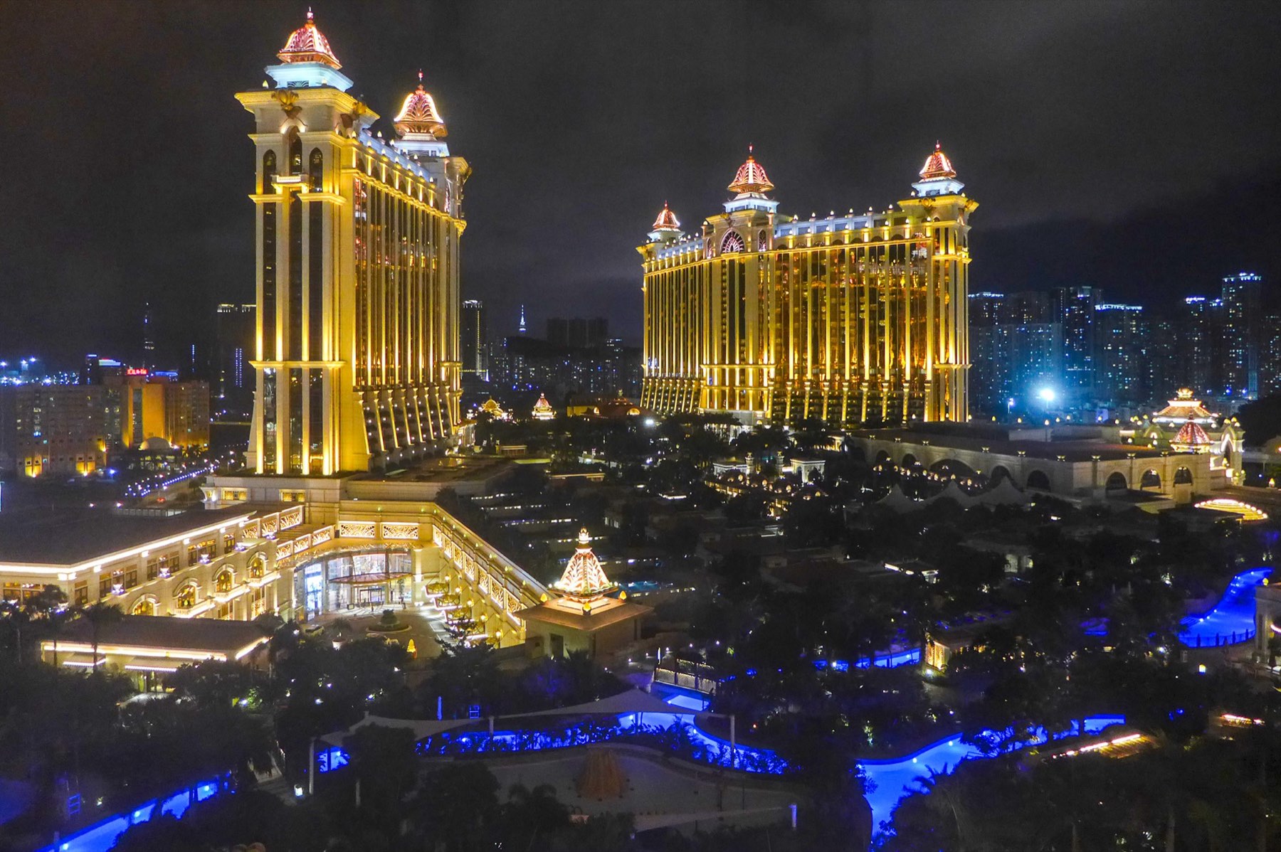 Galaxy Macau at night, its golden hotel towers with jewelled crowns lit above blue-lit pools and gardens in Cotai