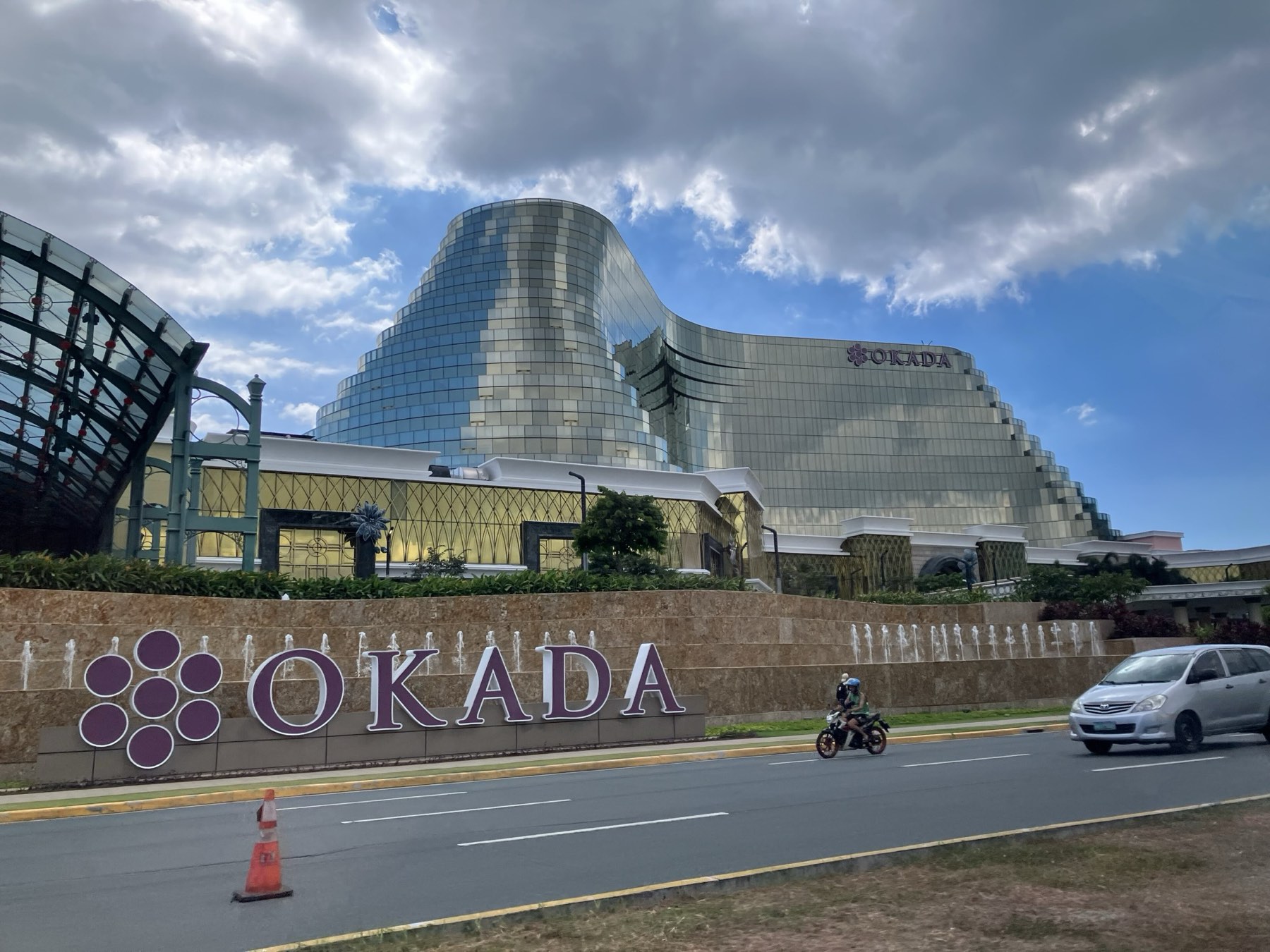 The curved gold-glass facade of Okada Manila behind a low stone wall with the purple Okada lettering and fountain jets